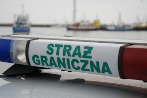 Fot. MOSG Napis Straż Graniczna na belce pojazdu służbowego, w tle kutry rybackie.